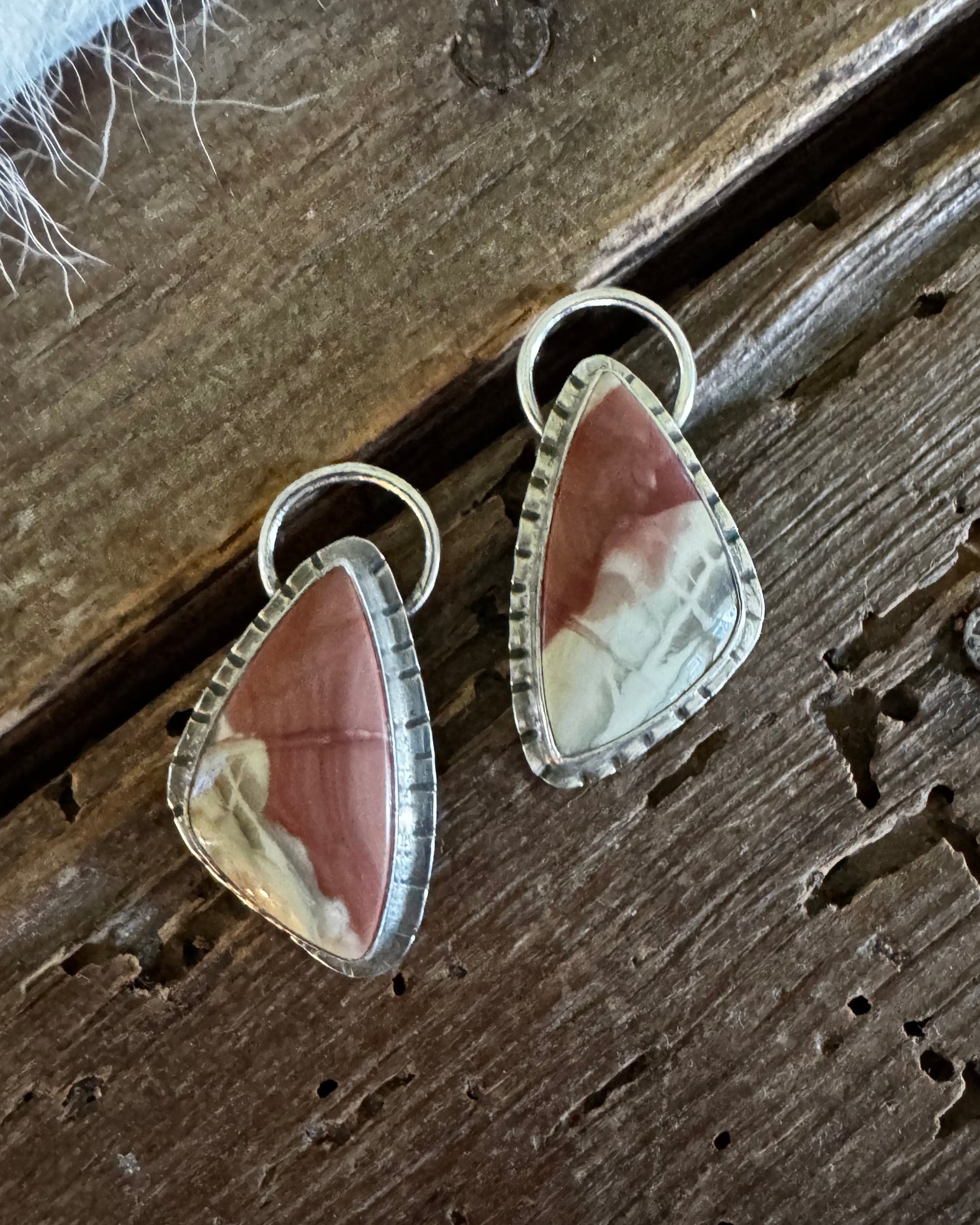 A pair of triangular drop earrings with pinkish-brown and cream marbled cabochon stones in silver bezel settings, displayed on a rustic dark wood surface.