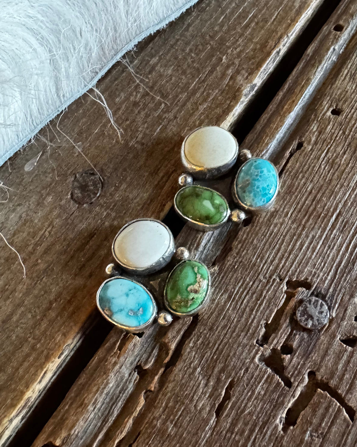 A pair of silver-toned stud earrings with oval turquoise-blue, green, and cream cabochon stones, displayed on a rustic wooden surface next to a white faux fur trim.
