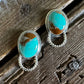 A pair of silver-toned drop earrings with oval turquoise and brown stone inlays, resting on a rustic wooden surface next to a white faux fur trim.