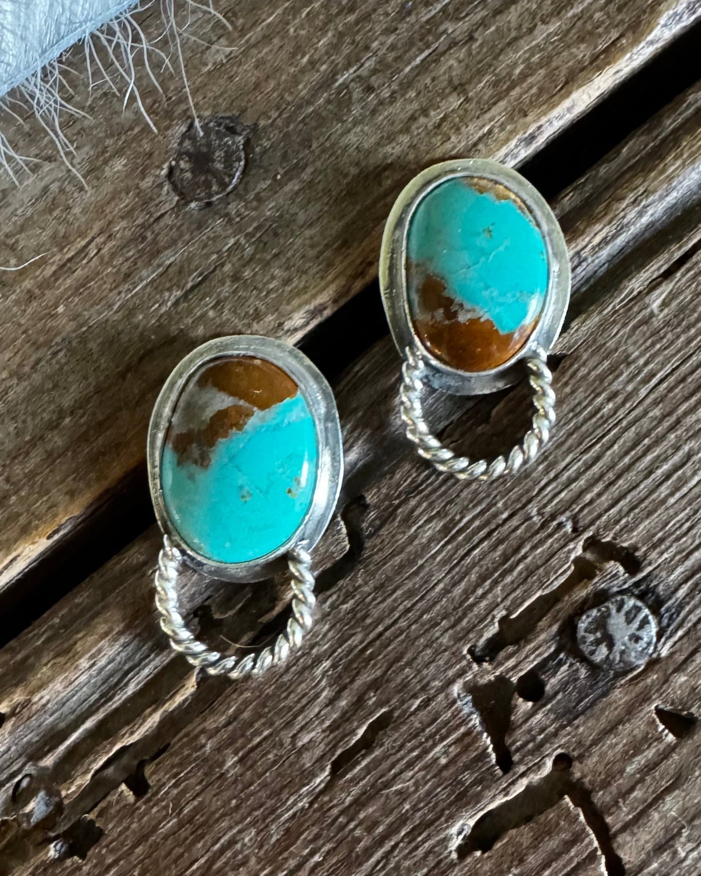 A pair of silver-toned drop earrings with oval turquoise and brown stone inlays, resting on a rustic wooden surface next to a white faux fur trim.