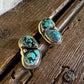 A pair of heart-shaped stud earrings with turquoise and black marbled stones in a gold setting, resting on a rustic wooden surface next to a white faux fur texture.
