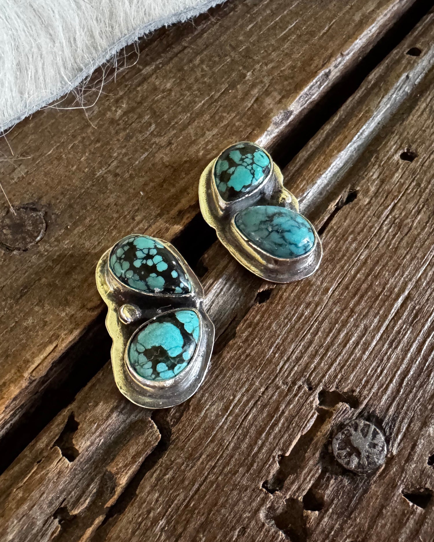 A pair of heart-shaped stud earrings with turquoise and black marbled stones in a gold setting, resting on a rustic wooden surface next to a white faux fur texture.