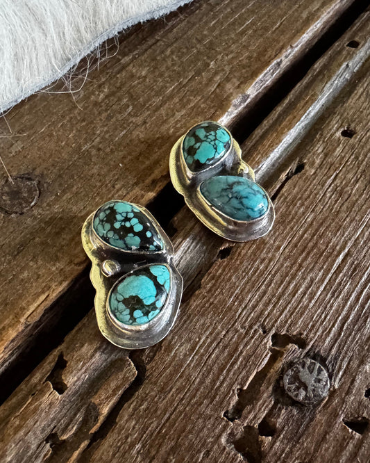 A pair of heart-shaped stud earrings with turquoise and black marbled stones in a gold setting, resting on a rustic wooden surface next to a white faux fur texture.