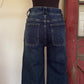 Back view of dark blue wide-leg jeans on a black dress form mannequin, set against a background with a cream wall and a rustic red wooden door.