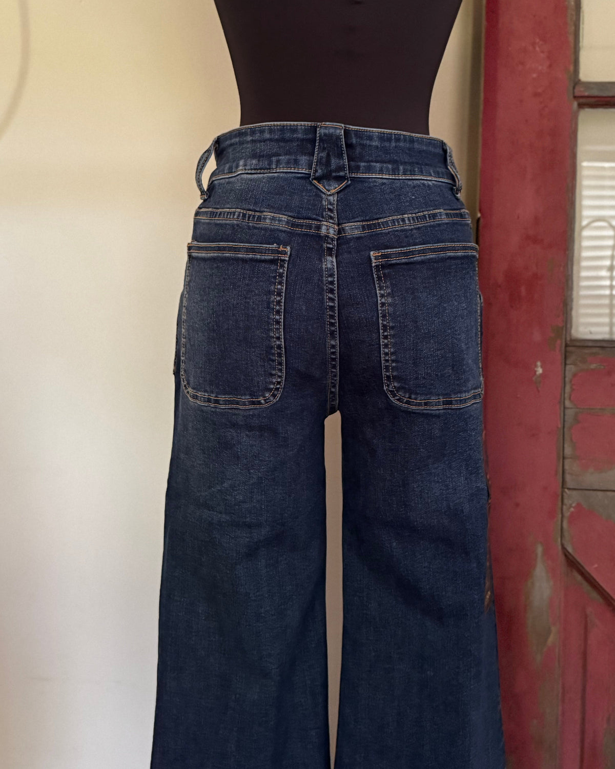 Back view of dark blue wide-leg jeans on a black dress form mannequin, set against a background with a cream wall and a rustic red wooden door.