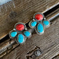 A pair of silver-toned stud earrings with red cabochon stones and turquoise inlay, displayed on a rustic wooden surface with a white faux-fur textile at the top.