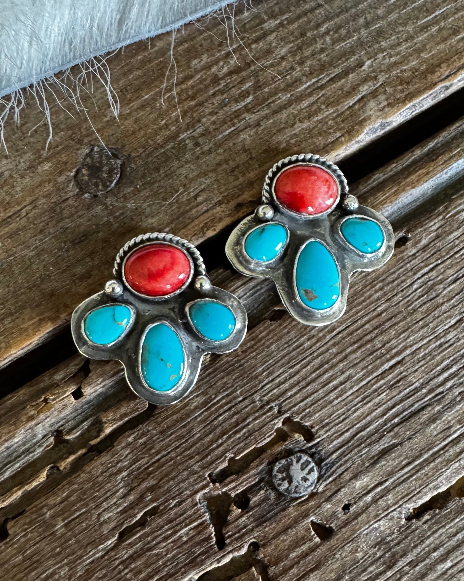 A pair of silver-toned stud earrings with red cabochon stones and turquoise inlay, displayed on a rustic wooden surface with a white faux-fur textile at the top.