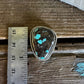 Turquoise stone in a gold frame on a wooden surface with a ruler for scale.
