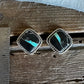 A pair of square silver-toned earrings with black inlay and turquoise-colored stone accents, displayed on a rustic wooden surface next to a metal ruler for scale.