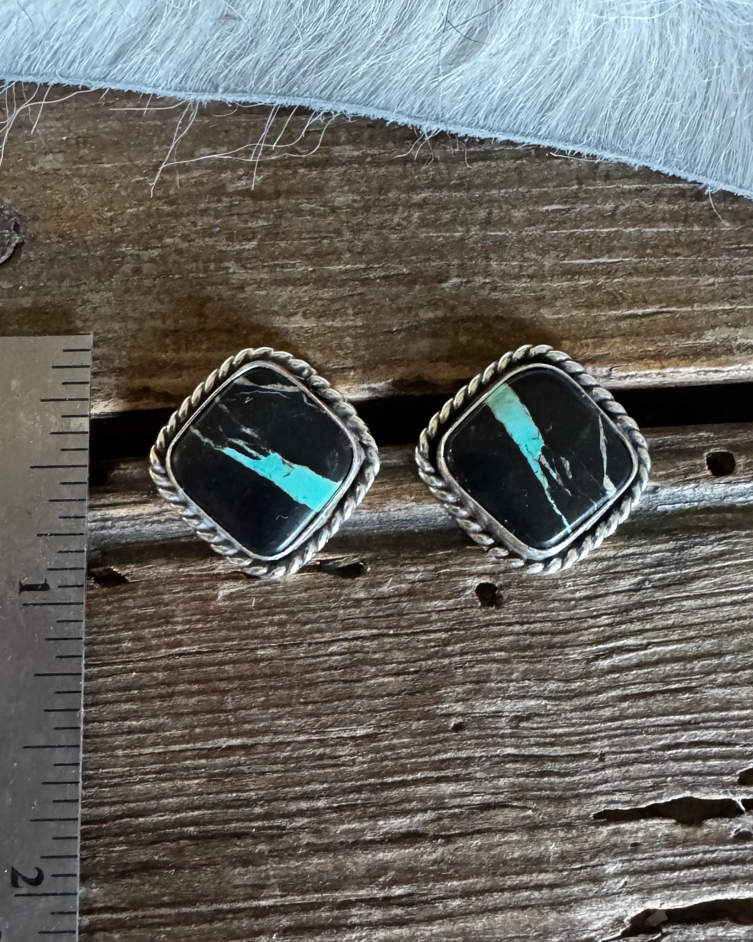 A pair of square silver-toned earrings with black inlay and turquoise-colored stone accents, displayed on a rustic wooden surface next to a metal ruler for scale.