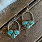 A pair of silver hoop earrings with three turquoise stones each, displayed on a rustic wooden surface next to a white faux fur trim.