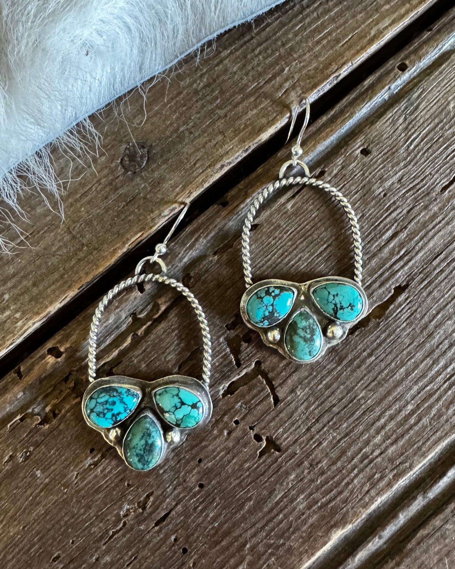 A pair of silver hoop earrings with three turquoise stones each, displayed on a rustic wooden surface next to a white faux fur trim.