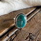 A silver ring with a round turquoise stone resting on a rustic wooden surface next to a white faux fur trim.