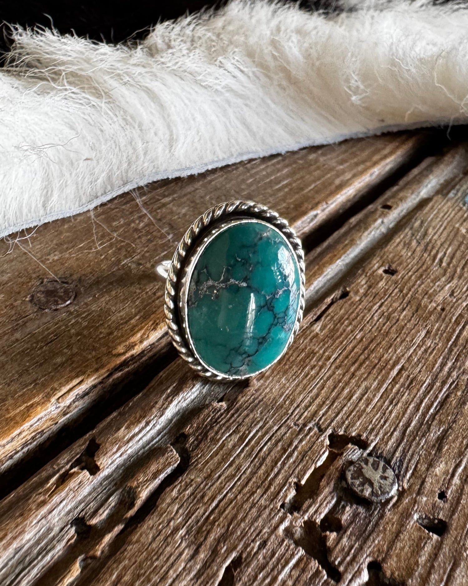 A silver ring with a round turquoise stone resting on a rustic wooden surface next to a white faux fur trim.