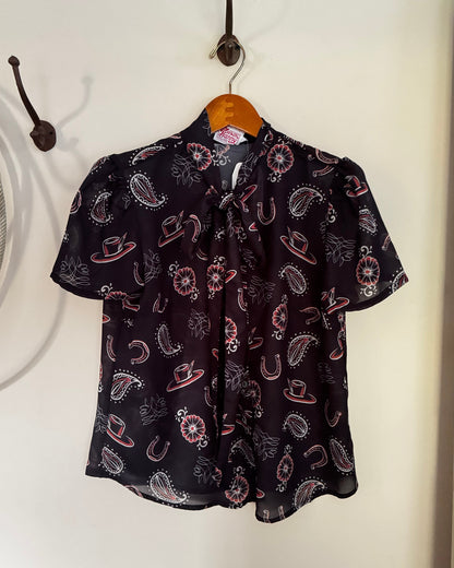 A short-sleeve button-up blouse with a western print of cowboy hats, horseshoes, and paisley motifs in pink, white, and gray on a black background, hanging on a wooden hanger from a wall hook.