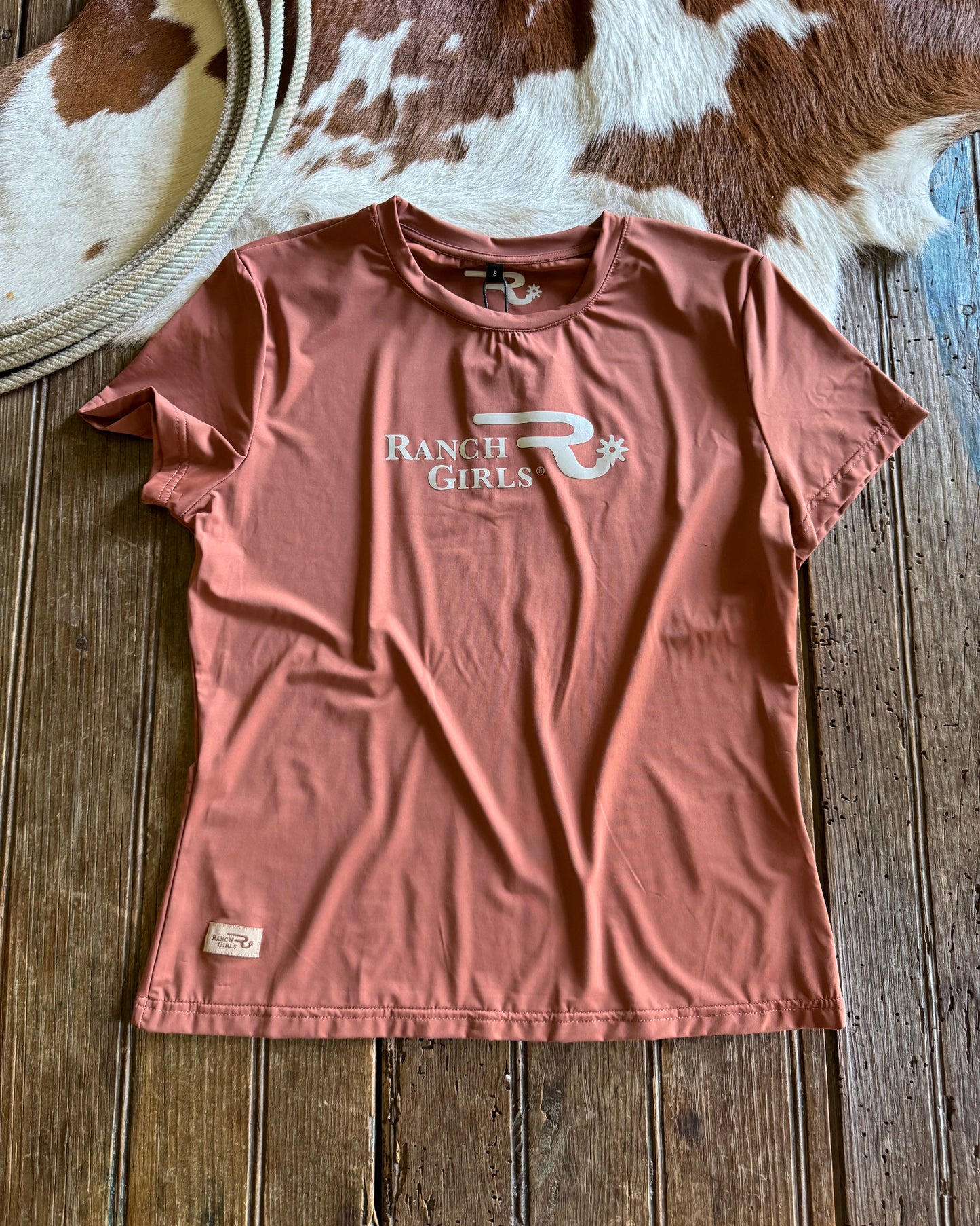 Pink t-shirt with 'Ranch Girls' logo on a wooden surface next to a cowhide rug