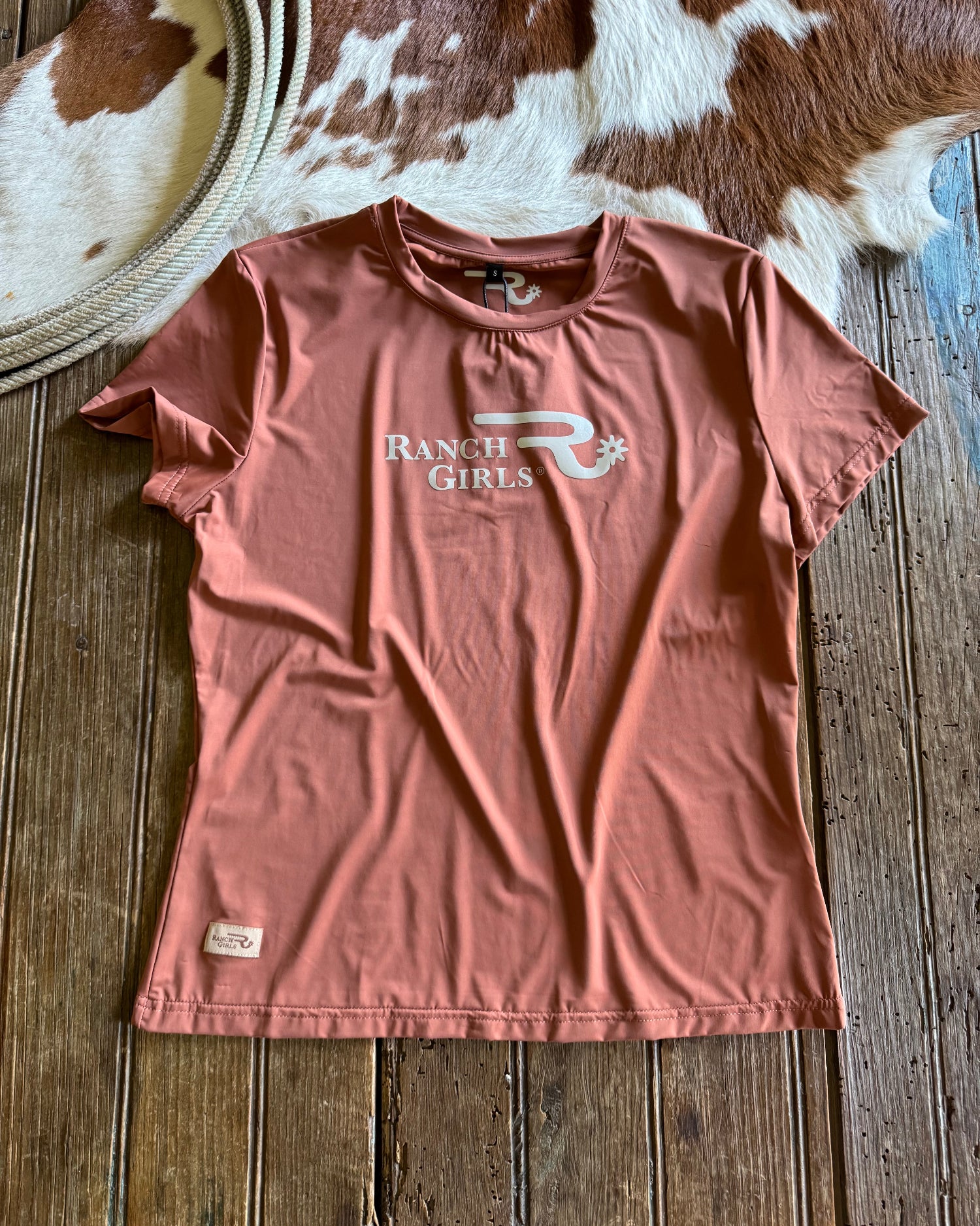 Pink t-shirt with 'Ranch Girls' logo on a wooden surface next to a cowhide rug