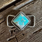 A silver twilly slide with a square turquoise stone set in a textured bezel, resting on a rustic dark brown wooden surface.