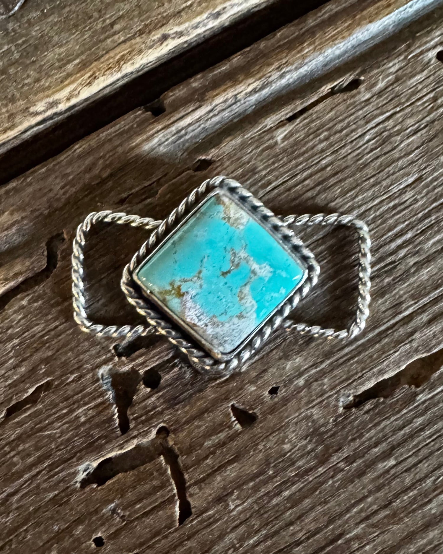 A silver twilly slide with a square turquoise stone set in a textured bezel, resting on a rustic dark brown wooden surface.