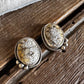 A pair of oval stud earrings with textured beige and brown stone centers and silver-toned bezel settings, resting on a rustic dark wood surface with white fur at the top.