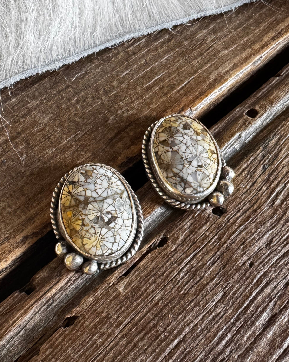 A pair of oval stud earrings with textured beige and brown stone centers and silver-toned bezel settings, resting on a rustic dark wood surface with white fur at the top.
