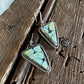 A pair of triangular turquoise-style drop earrings with silver wire-wrapped edges and fishhook backings, displayed on a rustic dark wood surface with a white faux-fur trim at the top left.