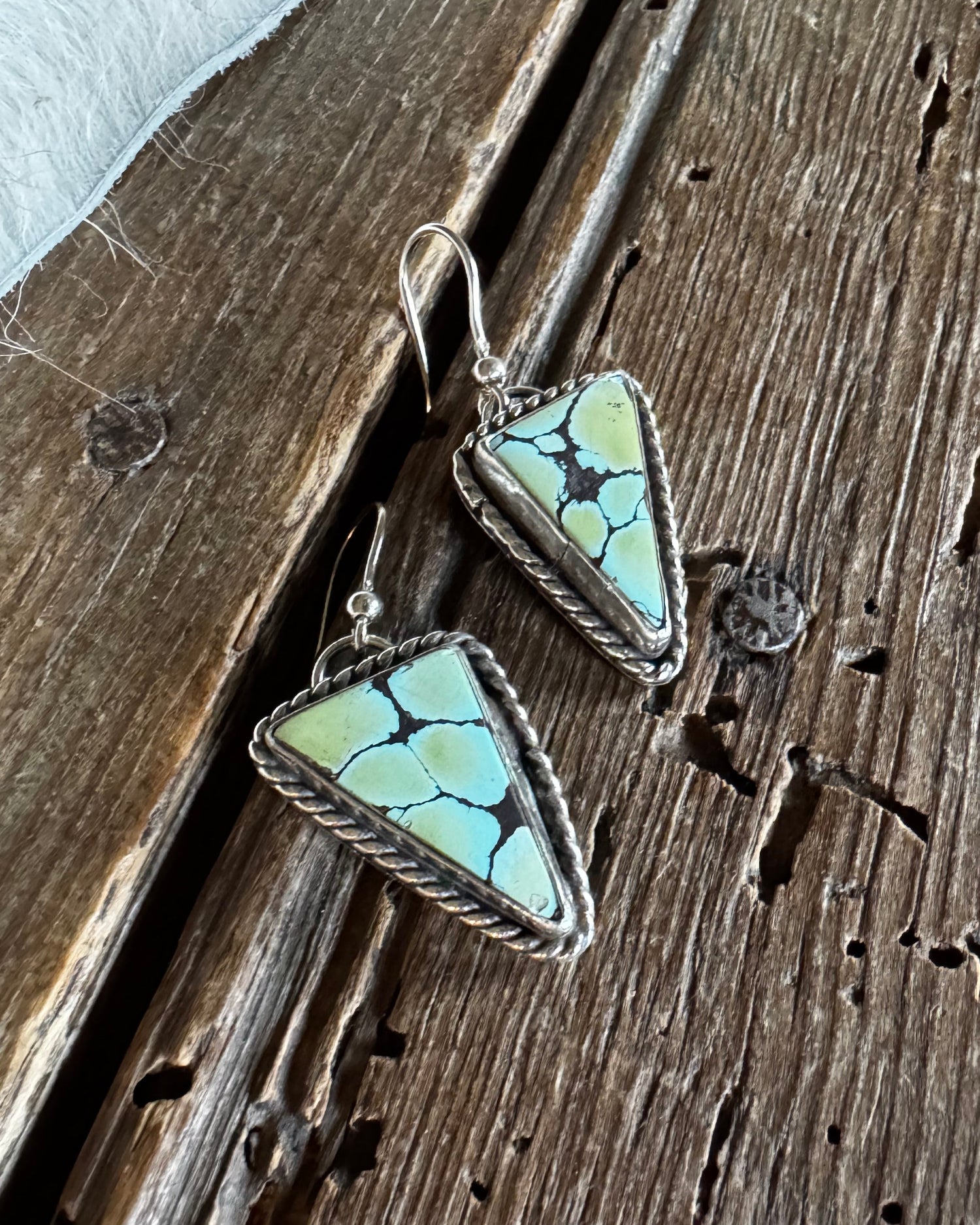 A pair of triangular turquoise-style drop earrings with silver wire-wrapped edges and fishhook backings, displayed on a rustic dark wood surface with a white faux-fur trim at the top left.