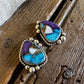 A pair of Southwestern-style turquoise and purple cabochon earrings with gold bezel settings, resting on a rustic wooden surface next to white faux fur.