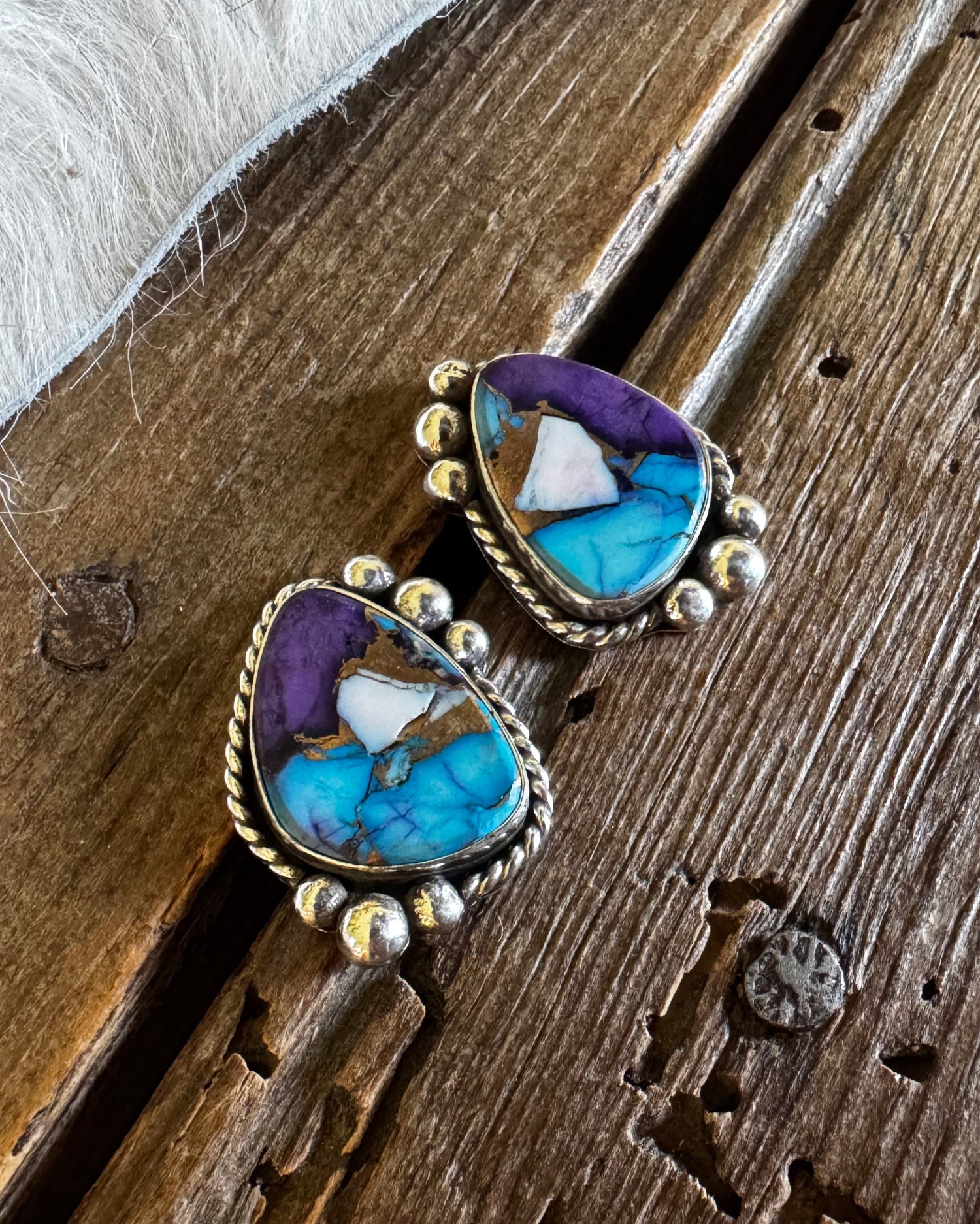A pair of Southwestern-style turquoise and purple cabochon earrings with gold bezel settings, resting on a rustic wooden surface next to white faux fur.