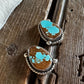 A pair of silver-toned rings with oval brown stones featuring bright turquoise blue matrix, displayed on a rustic dark wood surface next to a white fur trim.