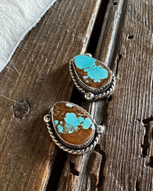 A pair of silver-toned rings with oval brown stones featuring bright turquoise blue matrix, displayed on a rustic dark wood surface next to a white fur trim.
