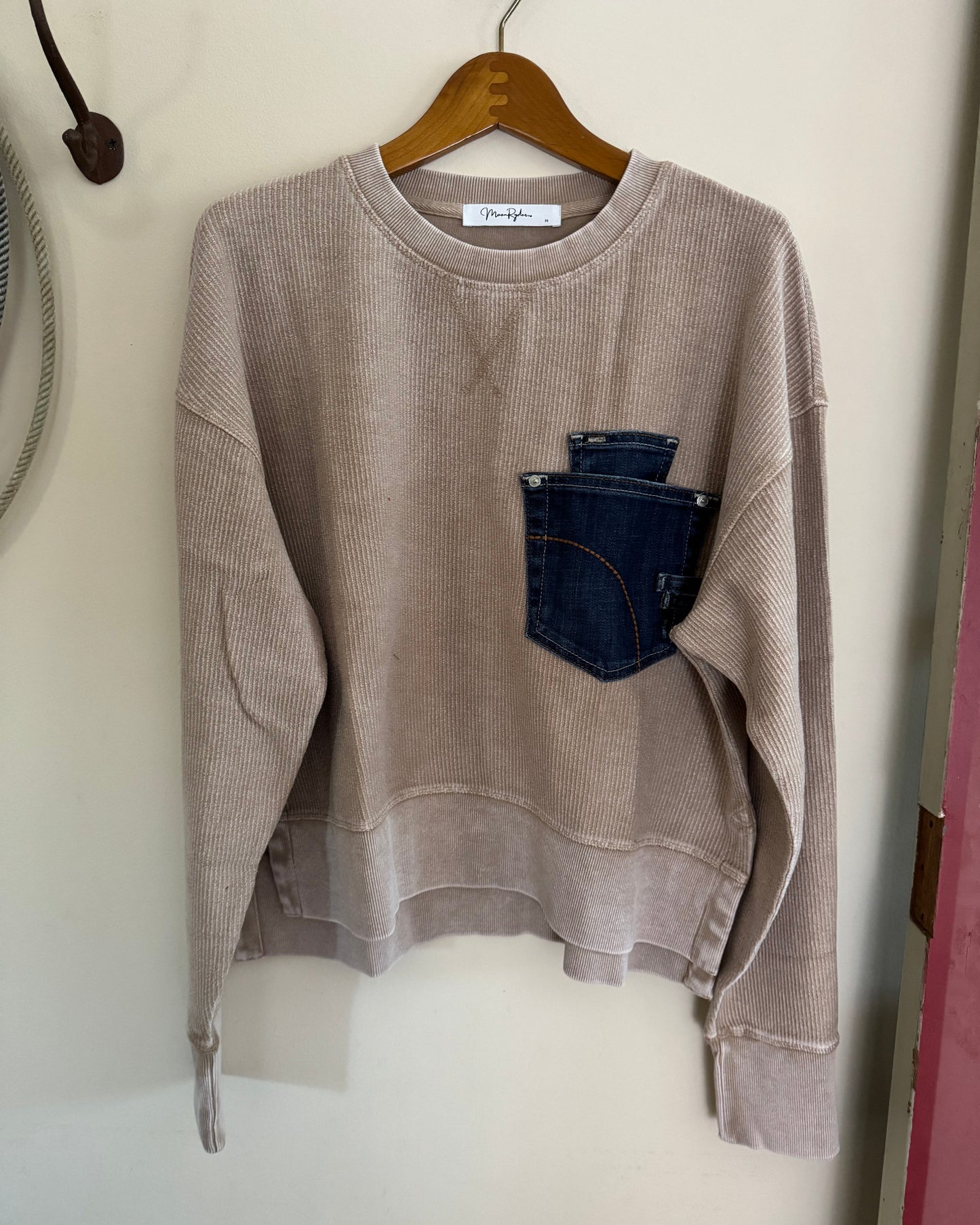 Beige sweatshirt with a denim pocket on a wooden hanger against a white wall.