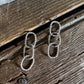 A pair of silver-toned drop earrings made of interlocking rope loops, displayed on a rustic dark wood surface with a white faux fur accent at the top.
