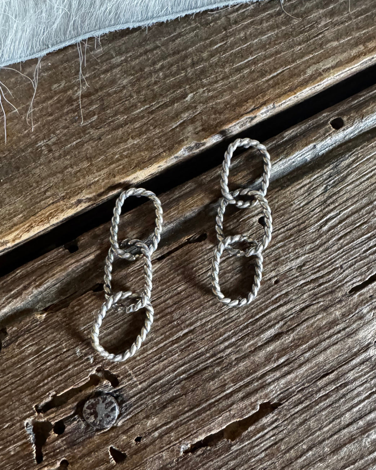 A pair of silver-toned drop earrings made of interlocking rope loops, displayed on a rustic dark wood surface with a white faux fur accent at the top.