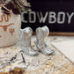 Silver cowboy boot earrings on a marble surface with a decorative sign in the background.
