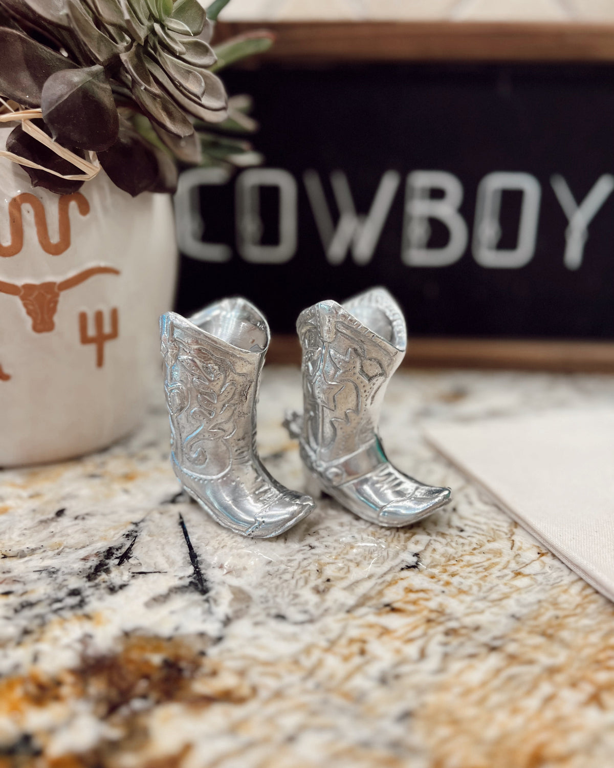 Silver cowboy boot earrings on a marble surface with a decorative sign in the background.