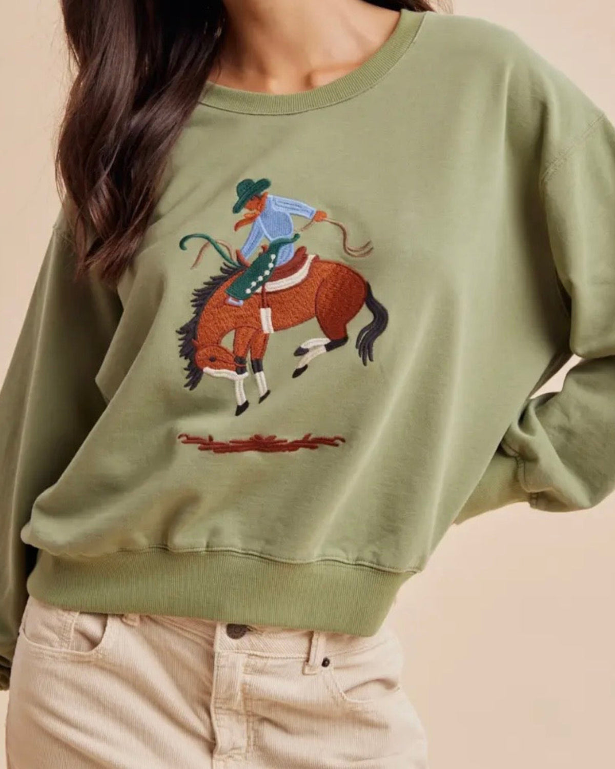 An olive green crewneck sweatshirt featuring a colorful embroidered graphic of a cowboy riding a horse, worn by a model with beige pants against a solid beige background.