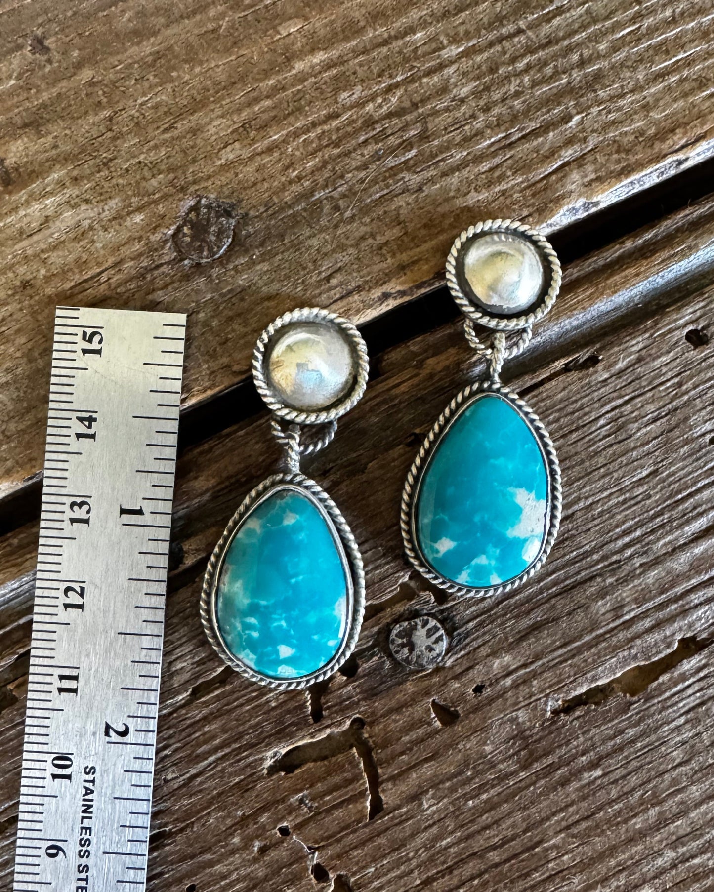 Pair of earrings with turquoise stones and pearl-like centers on a wooden surface with a ruler for scale.
