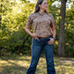 Woman wearing a patterned shirt and jeans standing in a natural setting with trees and grass.