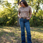 Woman standing in a natural setting wearing a patterned top and blue jeans.
