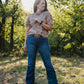 Woman wearing a patterned shirt and blue jeans standing in a forested area.