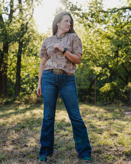 Woman wearing a patterned shirt and blue jeans standing in a forested area.