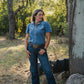 Woman in a blue shirt and jeans standing outdoors with trees and sunlight in the background