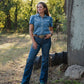 Woman in a blue denim outfit standing outdoors near a rustic building with trees in the background.