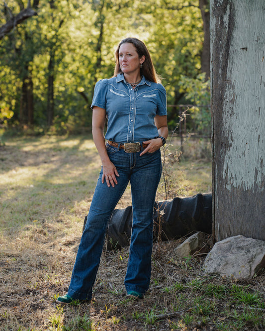 Woman in a blue denim outfit standing outdoors near a rustic building with trees in the background.