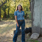 Woman in a denim outfit standing in a rural setting with trees and a wooden structure.