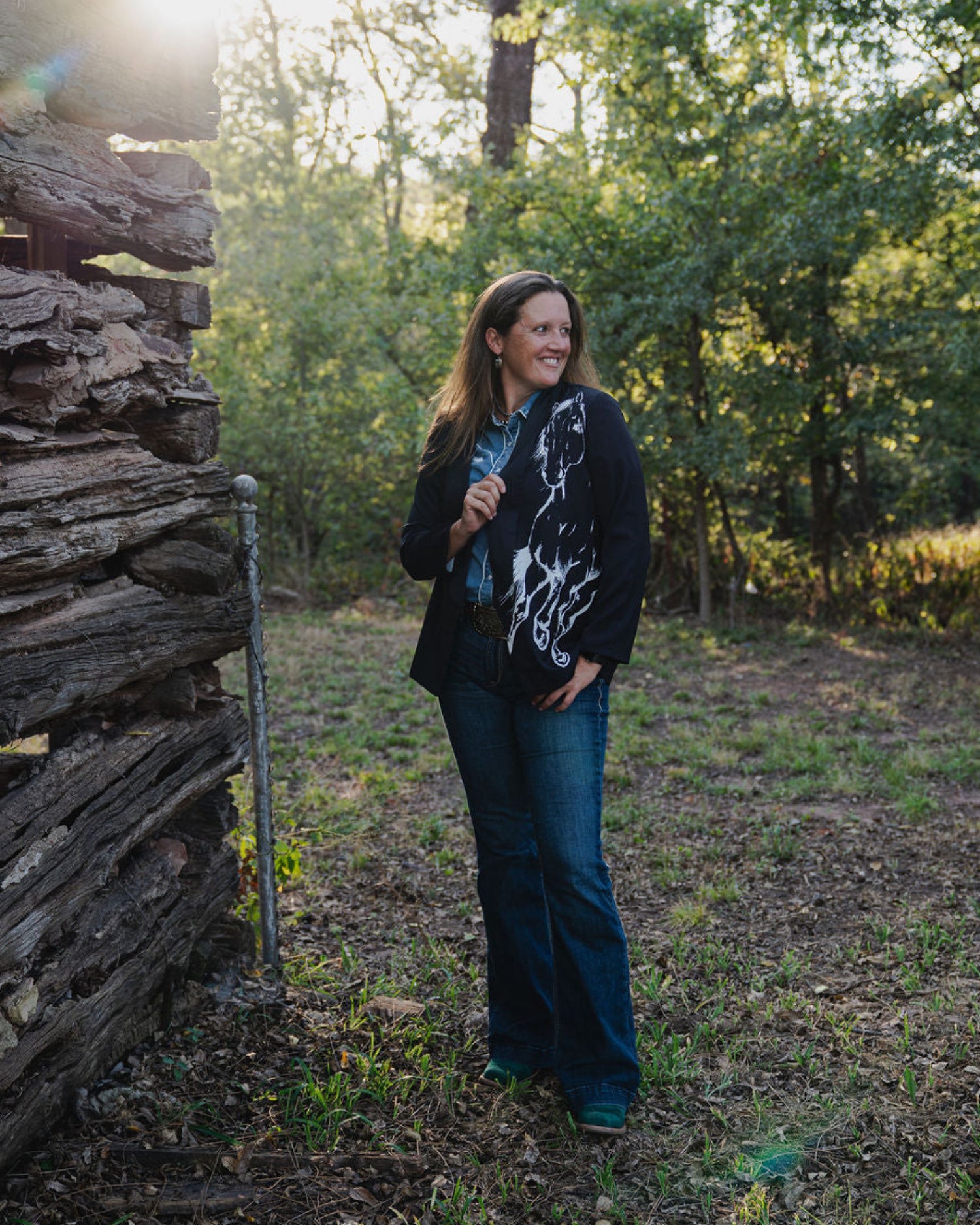 Woman standing in a forested area near a rustic wooden cabin.