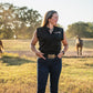 Woman standing in a field with horses, wearing a black shirt with a logo.