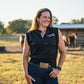 Woman wearing a black sleeveless shirt with a logo, standing in an outdoor setting with horses in the background.