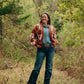 Woman standing in a forest wearing a patterned cardigan, denim jacket, and jeans.